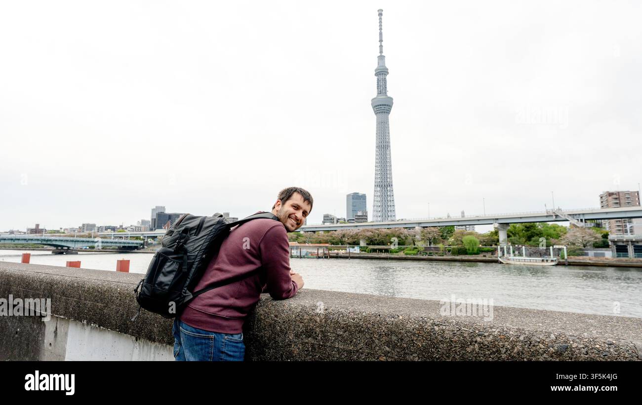 Viaggiatore sorridente con zaino si appoggia sull'argine del fiume Sumida a Tokyo, Giappone, con il Tokyo Skytree che si erge sopra il lungomare urbano e il ponte. Foto Stock
