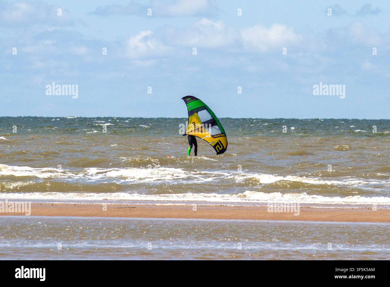 Un kitesurfer cavalca le onde agitate del mare vicino alla riva di una spiaggia sabbiosa sotto un cielo blu con nuvole sparse. Southport, UK. Foto Stock