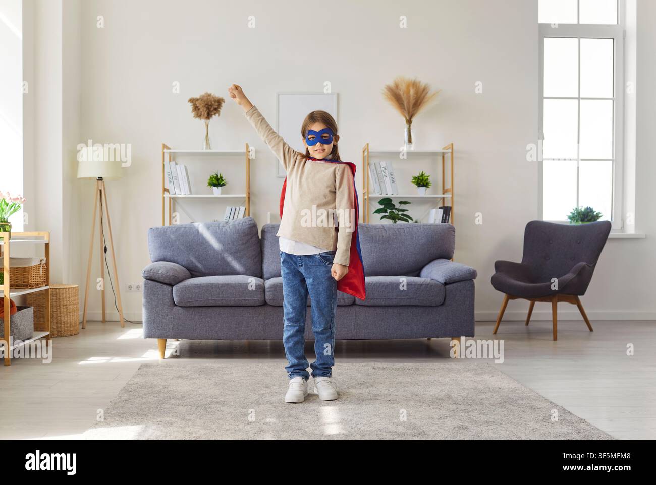 Ragazzo felice che indossa un costume da supereroe con mantello rosso e maschera blu mentre gioca a casa Foto Stock