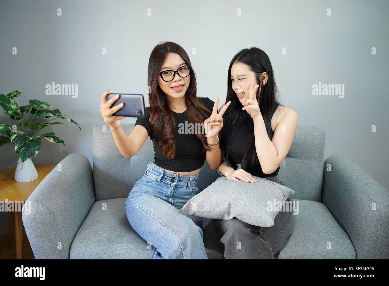Due giovani indonesiane sorridono e posano per un selfie con lo smartphone mentre si rilassano insieme su un divano a casa. Foto Stock