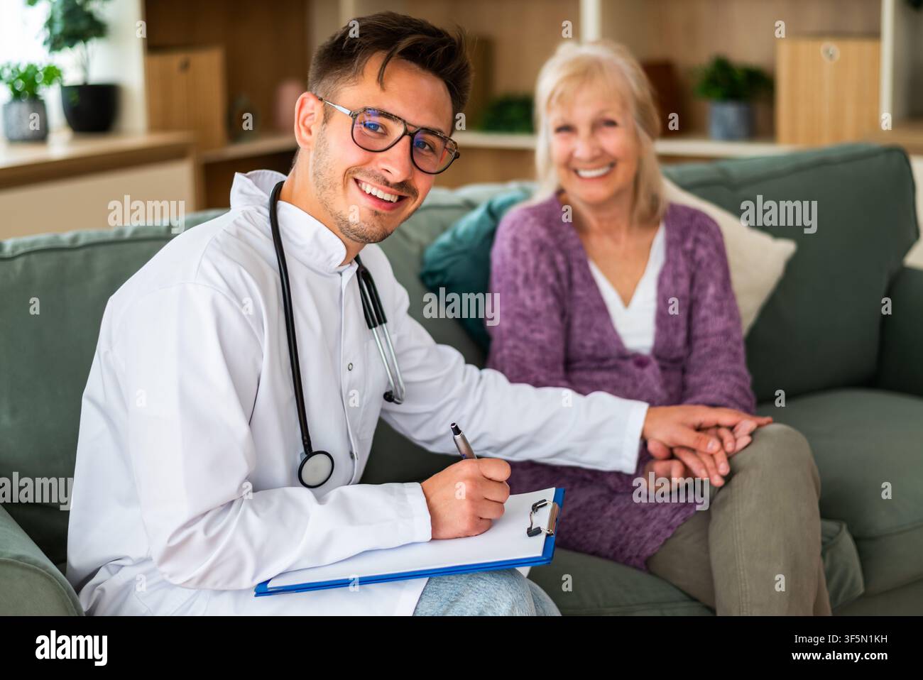 Un operatore sanitario cordiale sorride mentre prende appunti durante una visita a casa, tenendo delicatamente la mano di un paziente anziano per rassicurare e. Foto Stock