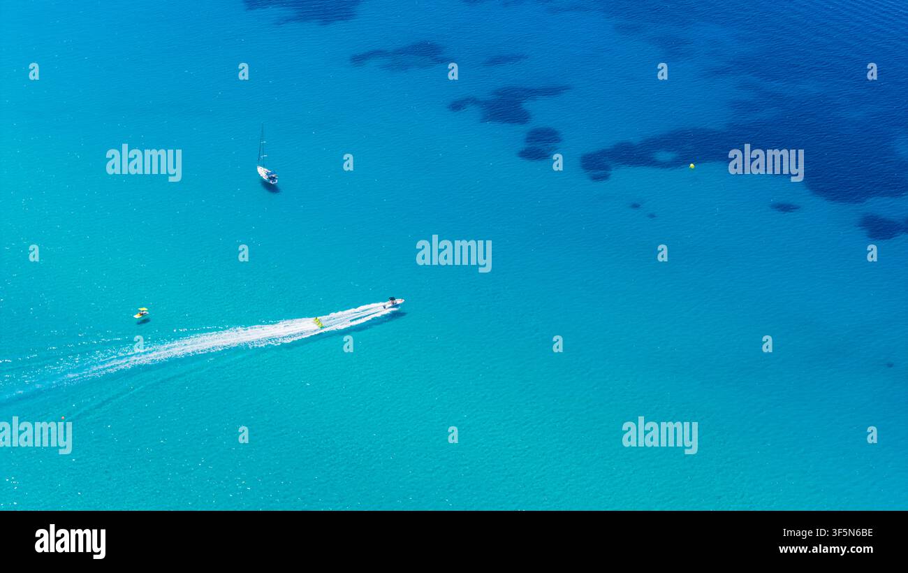 Acqua blu chiara circonda una barca bianca in rapido movimento e una piccola imbarcazione a vela, mentre macchie scure sott'acqua creano contrasto attraverso il paesaggio marino aperto Foto Stock