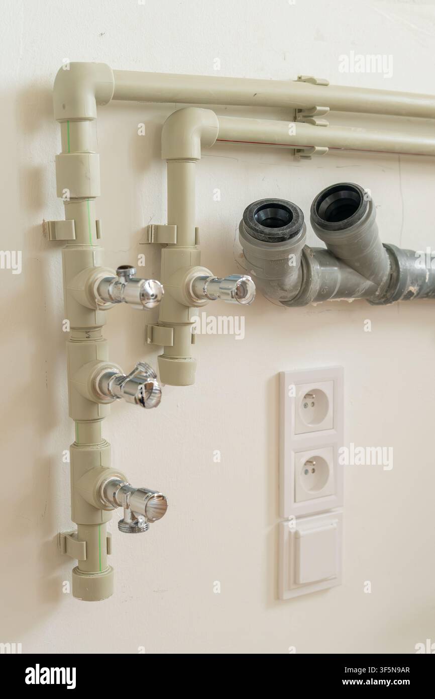 Sistema idraulico con rubinetti cromati collegati a tubi di plastica beige montati sulla parete. Tubi di scarico in PVC grigio visibili insieme a prese elettriche sullo sfondo. Foto Stock