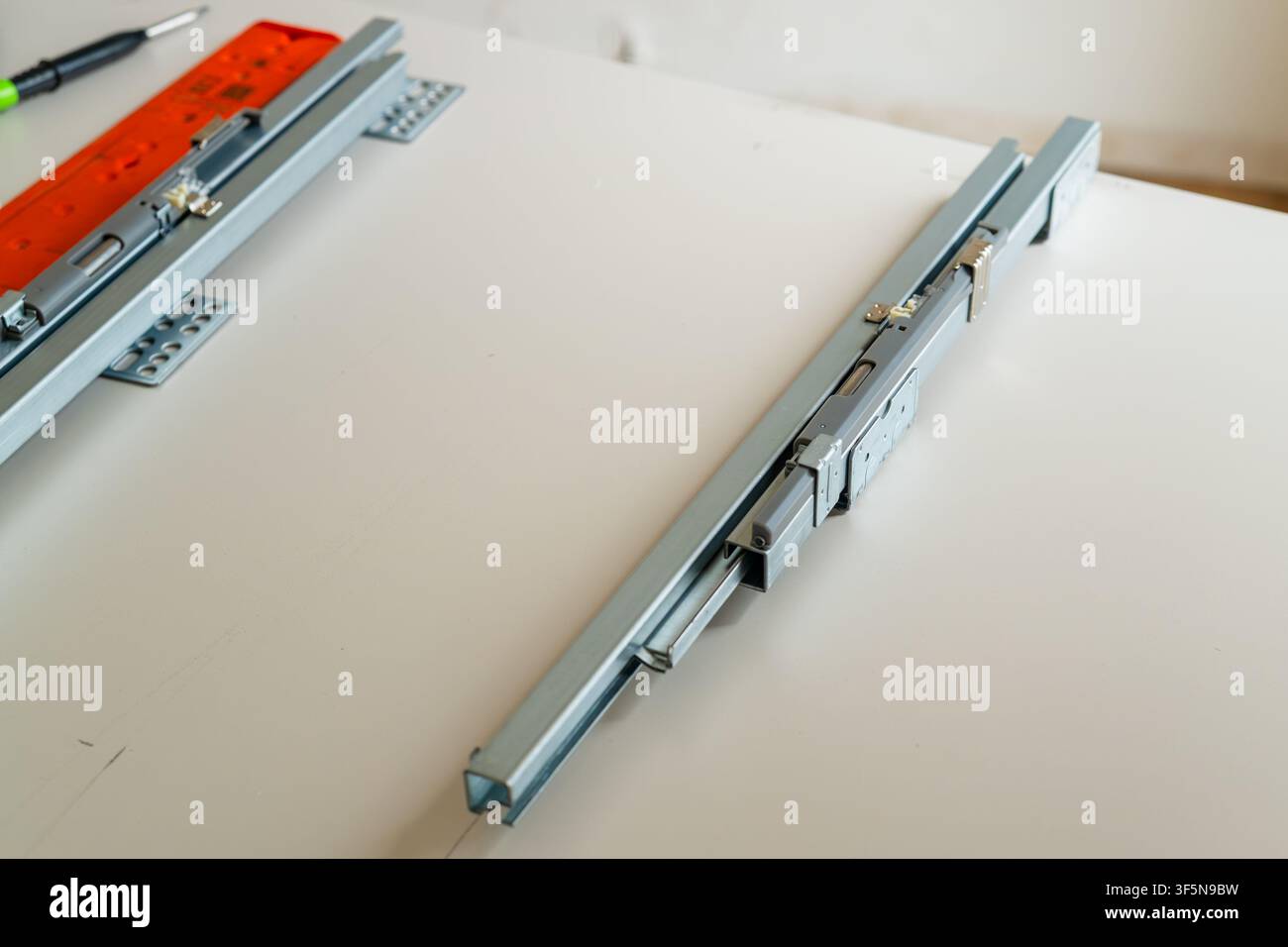 Installazione di guide con cuscinetti a sfere metallici su una superficie piana per l'assemblaggio del cassetto. I componenti visibili includono le guide, le viti e le staffe di fissaggio, con l'illuminazione ambientale che evidenzia le superfici metalliche. Foto Stock