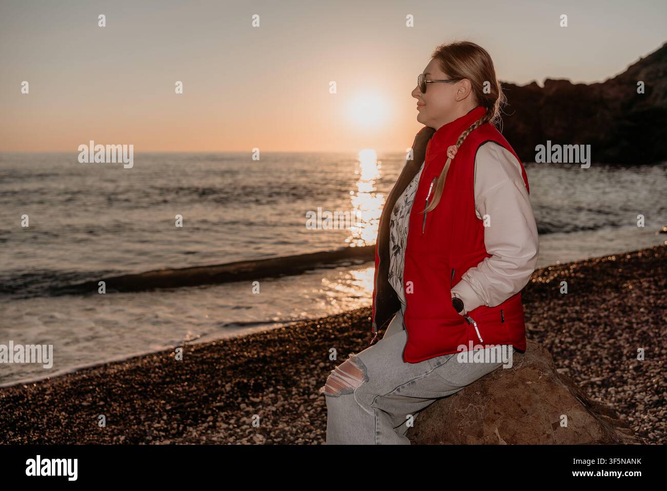 Donna tramonto spiaggia donna in giacca rossa seduta su una roccia al tramonto godendosi la vista scenica sull'oceano e la luce dorata dell'ora dorata Foto Stock