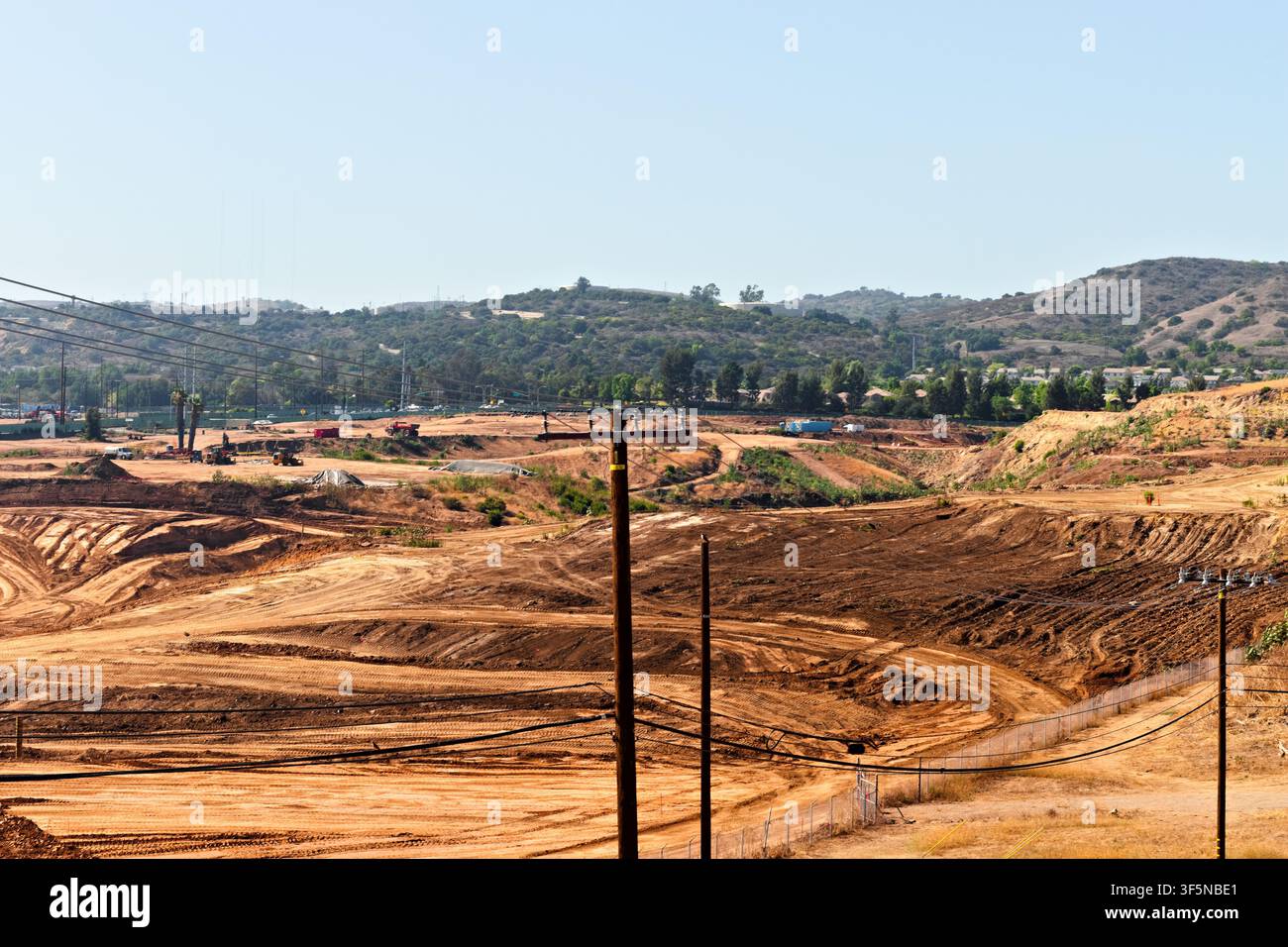 Un vasto cantiere edile si estende su colline aride, attraversato da tralicci e strade sterrate sinuose sotto un cielo estivo sereno. Foto Stock