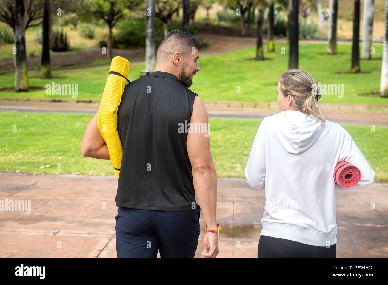 Due amici attivi che portano tappetini da yoga mentre camminano in un parco verde dopo aver terminato una sessione di fitness all'aperto. Foto Stock
