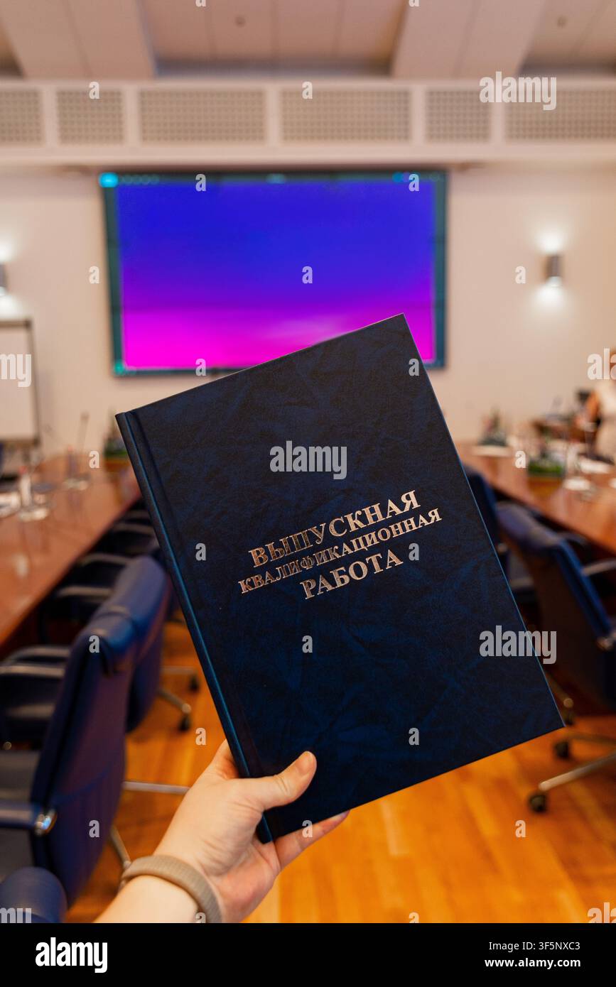 Mano tiene una copertina di tesi blu scuro in una sala conferenze. Il titolo recita Tesi di laurea. La stanza ha tavoli e sedie di legno. Un grande schermo mostra un gradie viola Foto Stock
