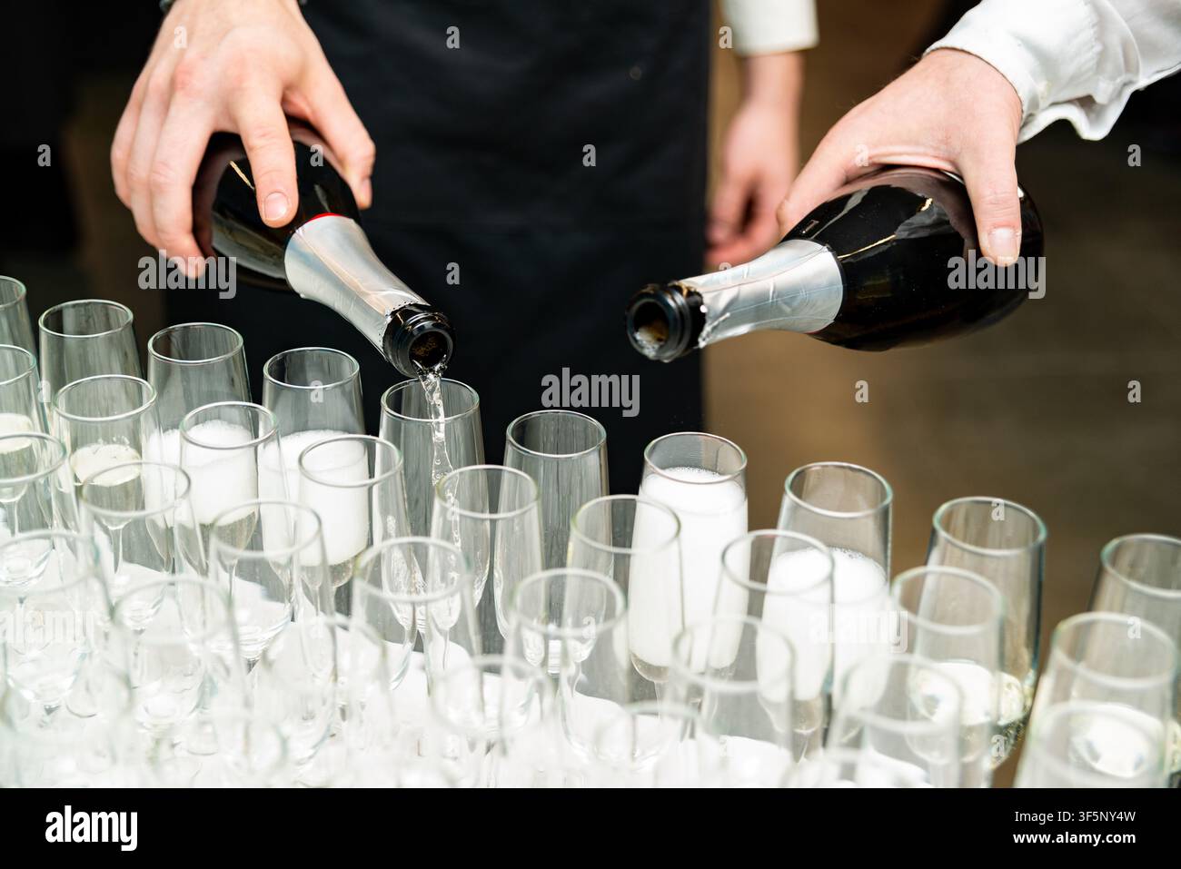 Si vedono le mani di due individui che riempiono file di calici di champagne vuoti e parzialmente riempiti con vino spumante, suggerendo la preparazione di una celebrazione Foto Stock