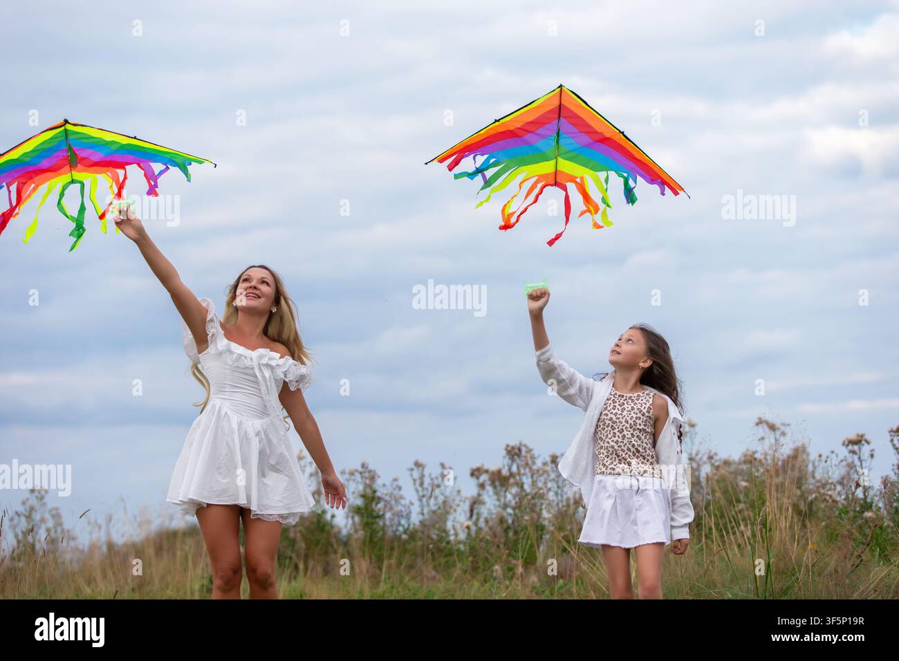 Una scena gioiosa mostra una donna e una figlia che volano sui serpenti luminosi dell'arcobaleno in una giornata nuvolosa. Si trovano sul campo erboso, godendosi la mo Foto Stock