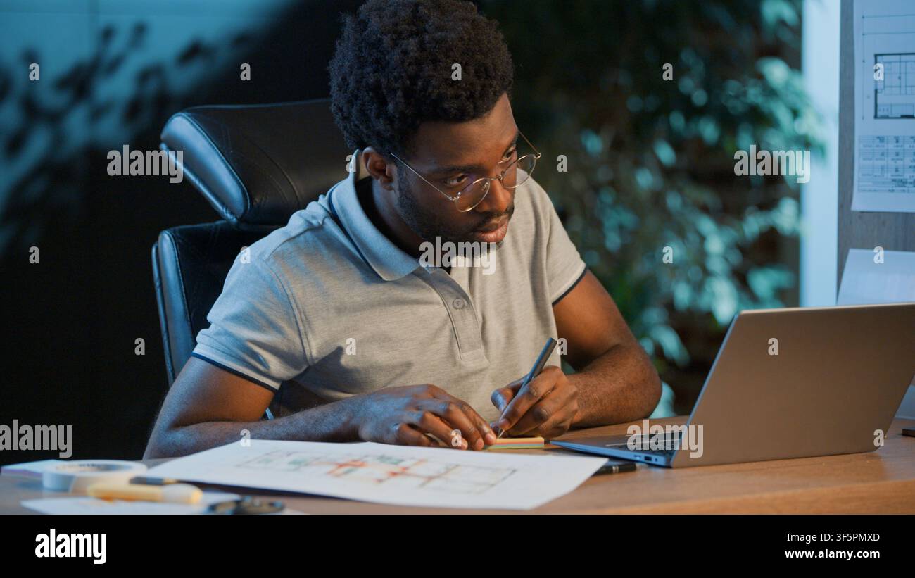 Uomo d'affari afroamericano concentrato, architetto che analizza progetti di costruzione e scrive appunti con un laptop in un ufficio notturno, uomo concentrato Foto Stock