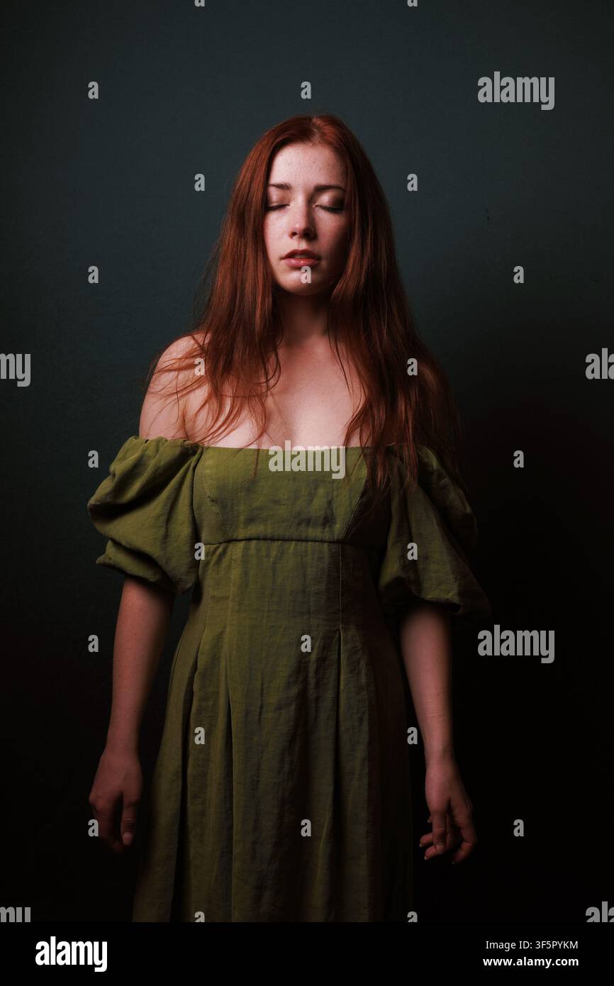 Donna dai capelli rossi in vestito verde oliva con gli occhi chiusi che evoca calma, introspezione e serenità silenziosa Foto Stock