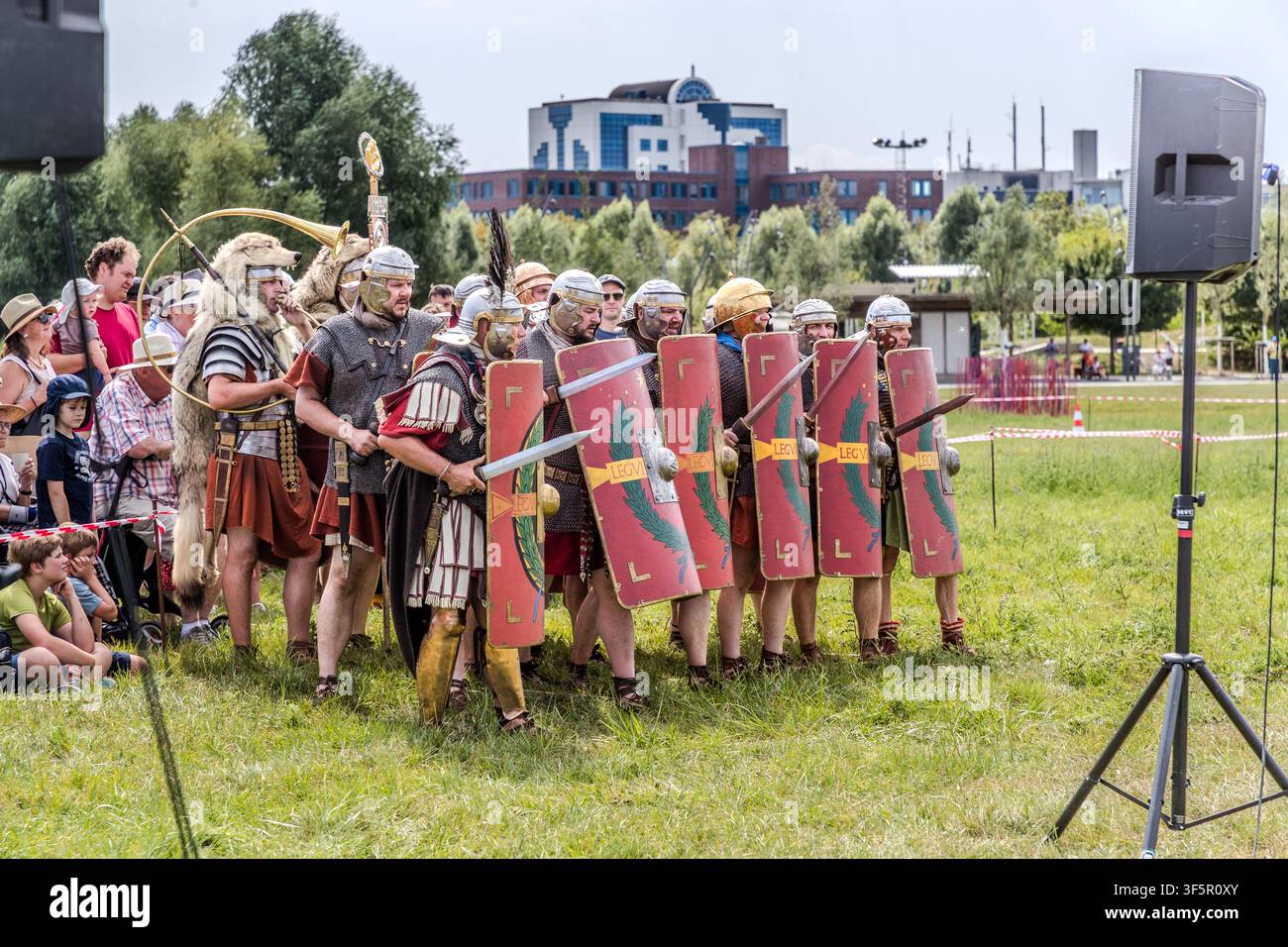 I legionari della Legio VI Victrix si trovano in fila con le spade sguainate, un trombettiere con un cappuccio di pelle di lupo e un centurione adornato di medaglie phalerae accompagnano la formazione a sinistra. Gli edifici moderni della Landesgartenschau (LAGA) a Neuss, Germania, si ergono a destra sullo sfondo. Foto Stock