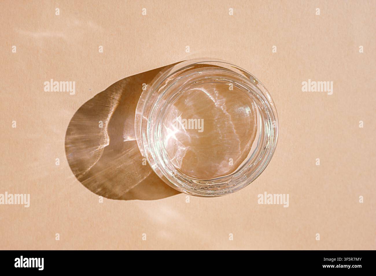 Vista dall'alto di un bicchiere d'acqua limpida che proietta riflessi di luce solare e un'ombra morbida su carta beige Foto Stock
