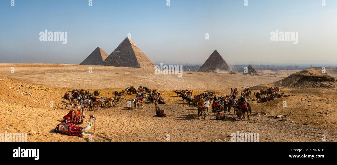 Vista della necropoli di Giza con cammelli e cavalieri che riposano sulla sabbia del deserto sotto un cielo sereno ad Al Haram, governatorato di Giza, Egitto. Foto Stock