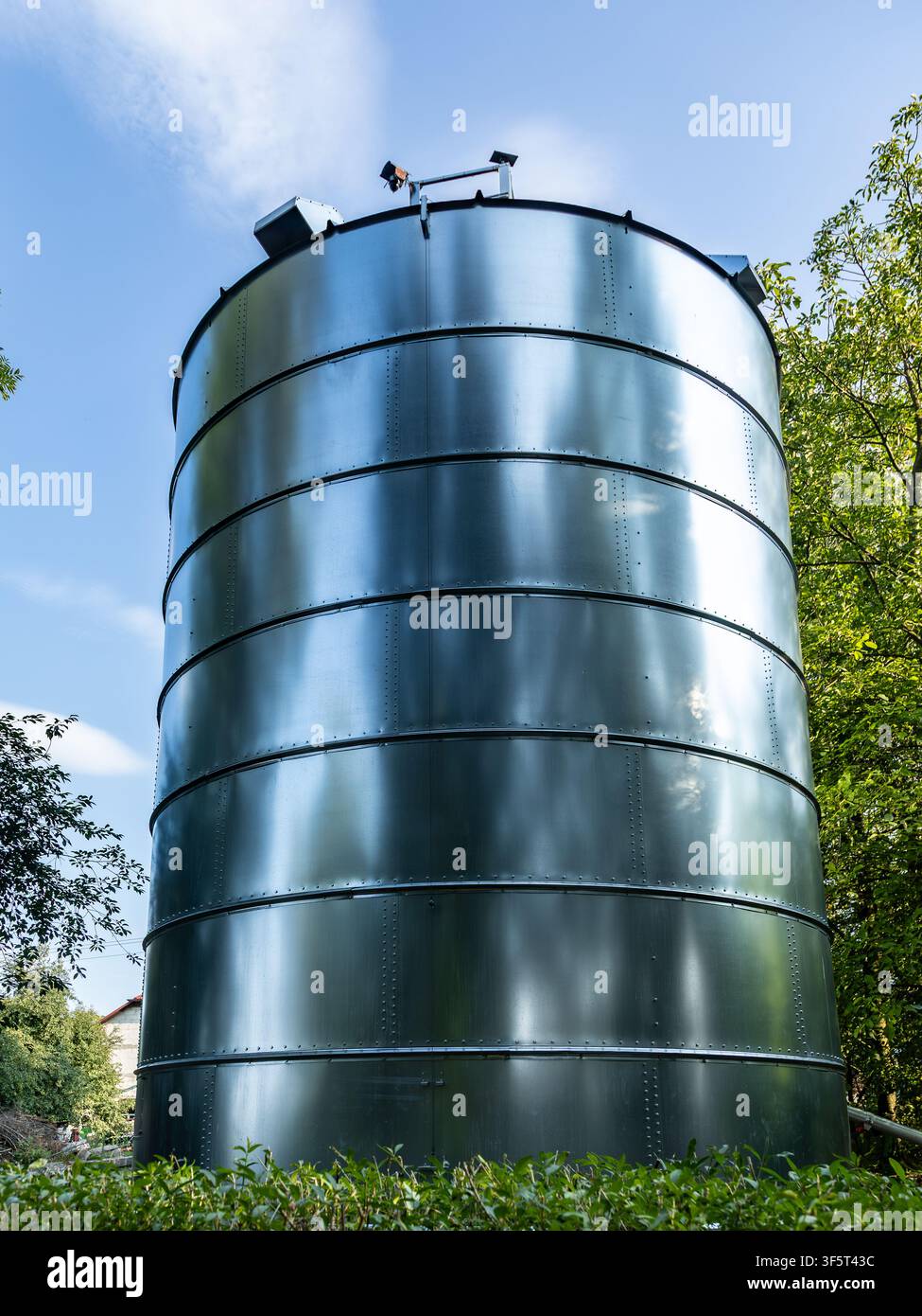 Serbatoio cilindrico industriale circondato dalla vegetazione sotto un cielo azzurro estivo. Foto Stock