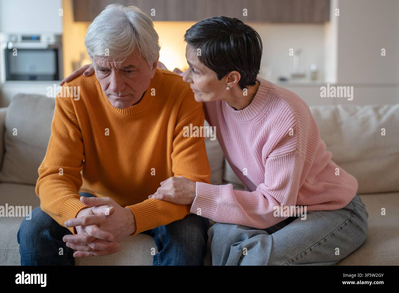 Coppia di anziani seduti sul divano a casa, una donna anziana espressiva consola il marito sopraffatto e sconvolto. Foto Stock
