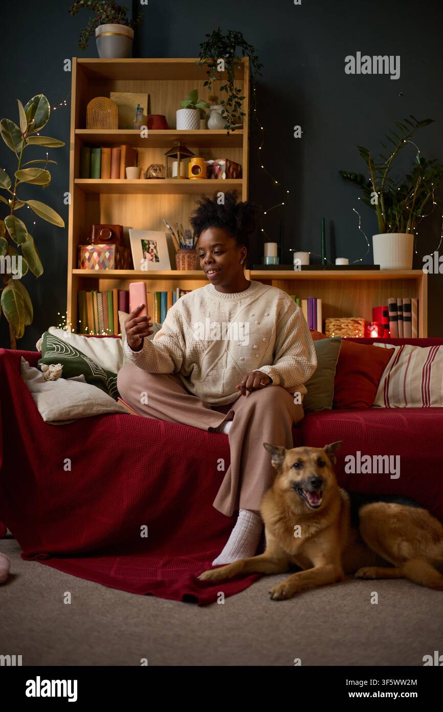 Immagine verticale di una giovane donna afroamericana seduta su un divano e che tiene uno smartphone durante una videochiamata, cane sdraiato sul pavimento, rappresentante la comunicazione remota e la proprietà di animali domestici Foto Stock