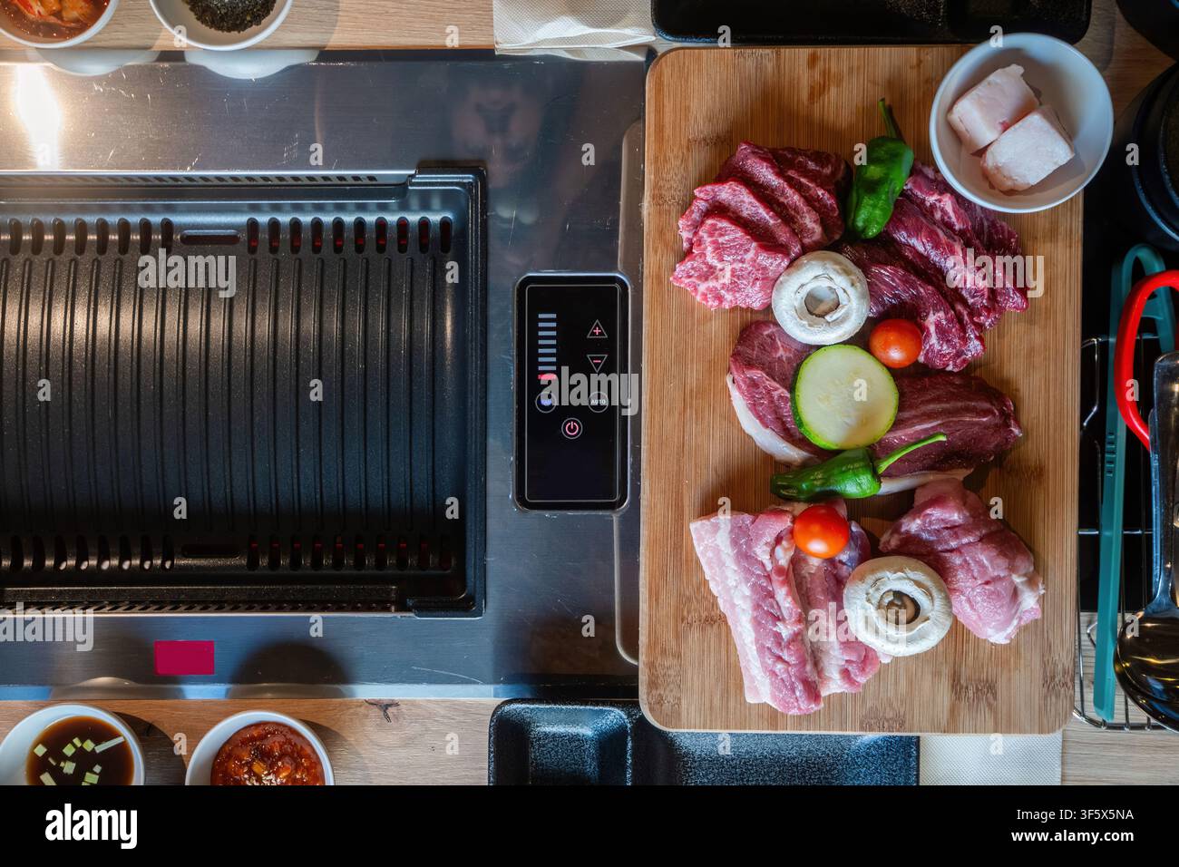 Tagli di maiale crudi disposti su una griglia elettrica da tavolo nera per barbecue coreano, visti direttamente dall'alto Foto Stock