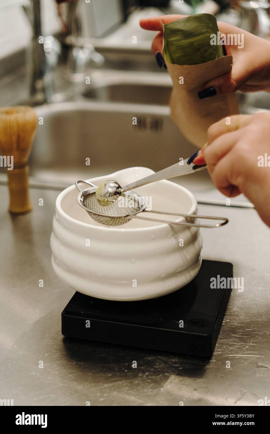 Mani che misurano polvere di matcha premium attraverso un colino fine in una ciotola di ceramica. Preparazione tradizionale del tè giapponese con strumenti di precisione su un Foto Stock