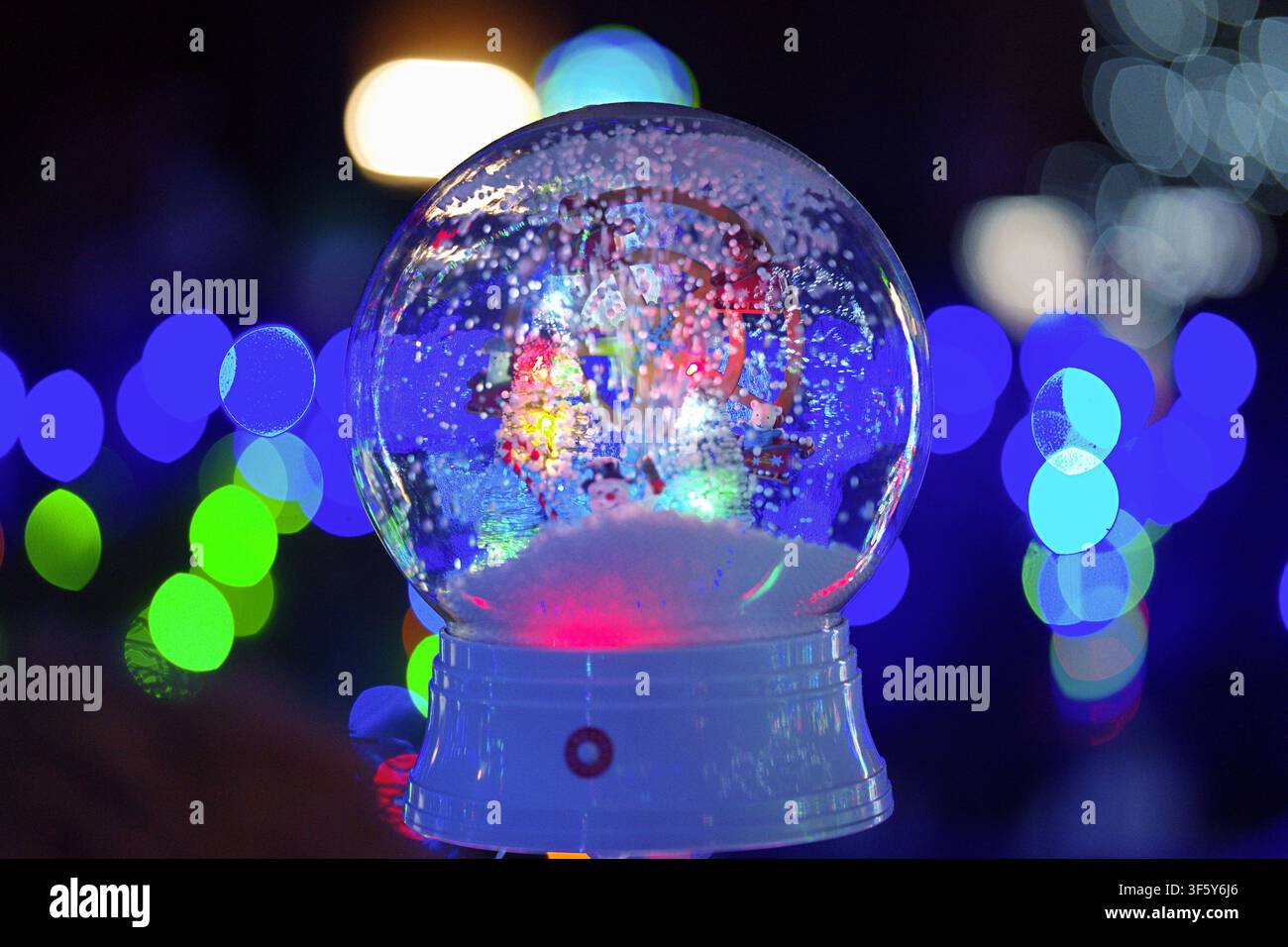 Un primo piano di una palla di neve che gira di notte, adornata con luci bokeh, su uno sfondo bianco e rosso, decorazioni natalizie e celebrazioni festive. Foto Stock