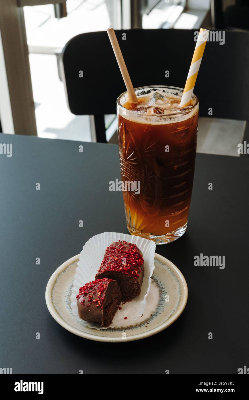 Caffè freddo rinfrescante servito con un dessert ricoperto di cioccolato guarnito con pezzi di lampone liofilizzati. Tavolo da caffè moderno con minimalismo Foto Stock