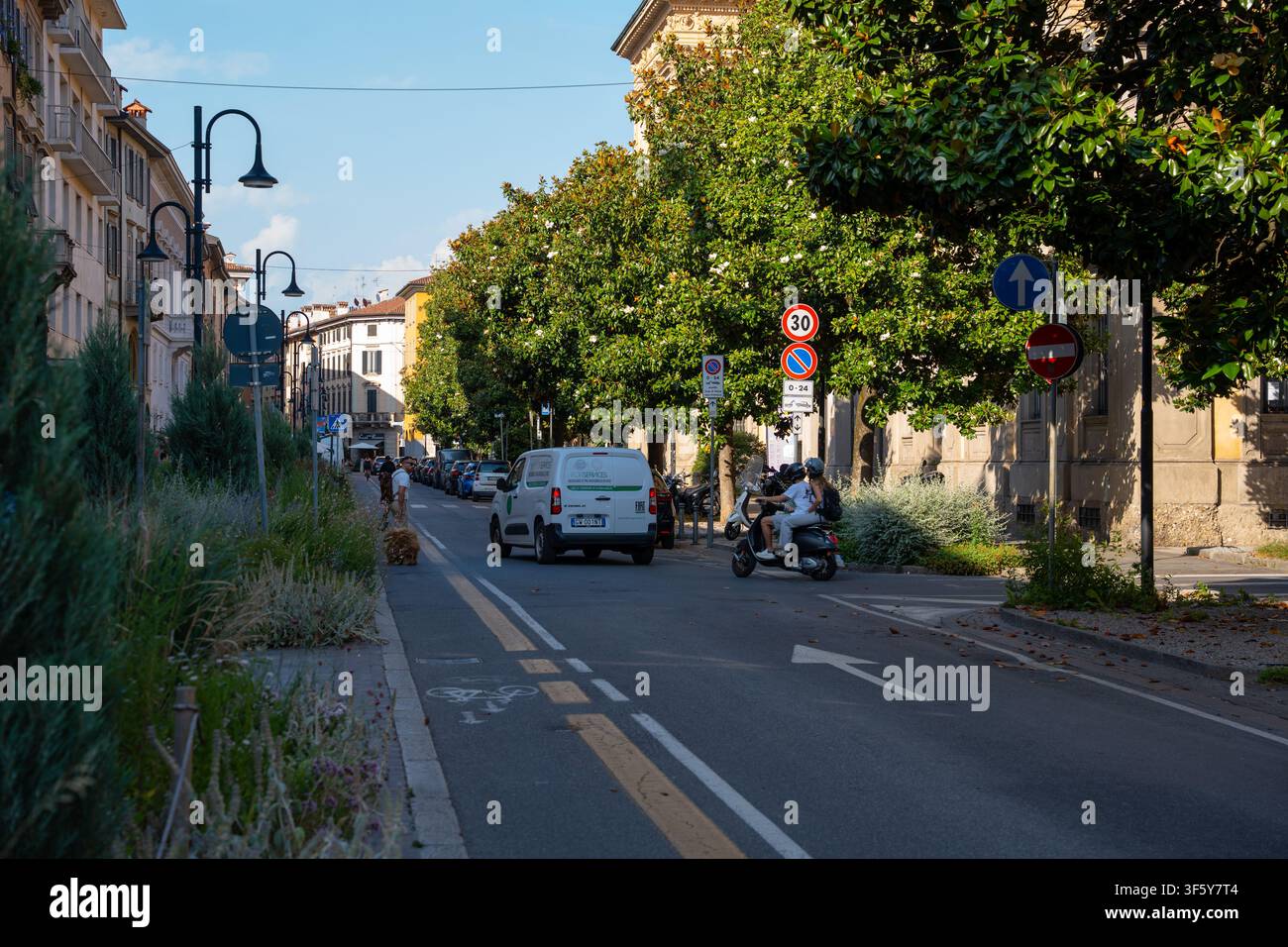 Bergamo, Italia – 16 giugno 2026: Foto dinamica del traffico urbano che mostra un furgone per le consegne e uno scooter che percorrono una strada cittadina italiana fiancheggiata da una vegetazione lussureggiante Foto Stock