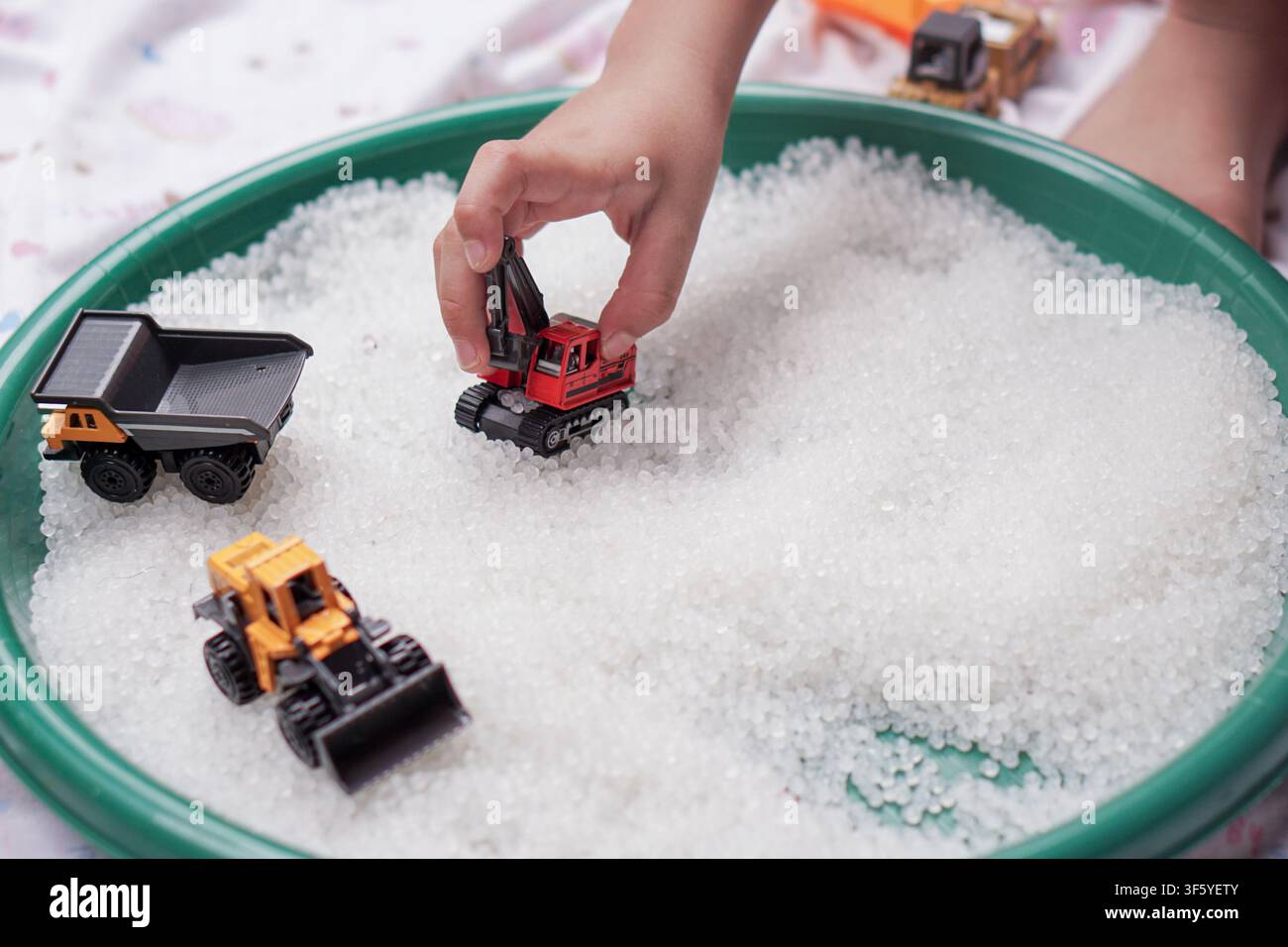 Mano di un bambino che utilizza un escavatore giocattolo e camion in un vassoio sensoriale pieno di perline per il gioco di sviluppo e l'apprendimento tattile. Foto Stock