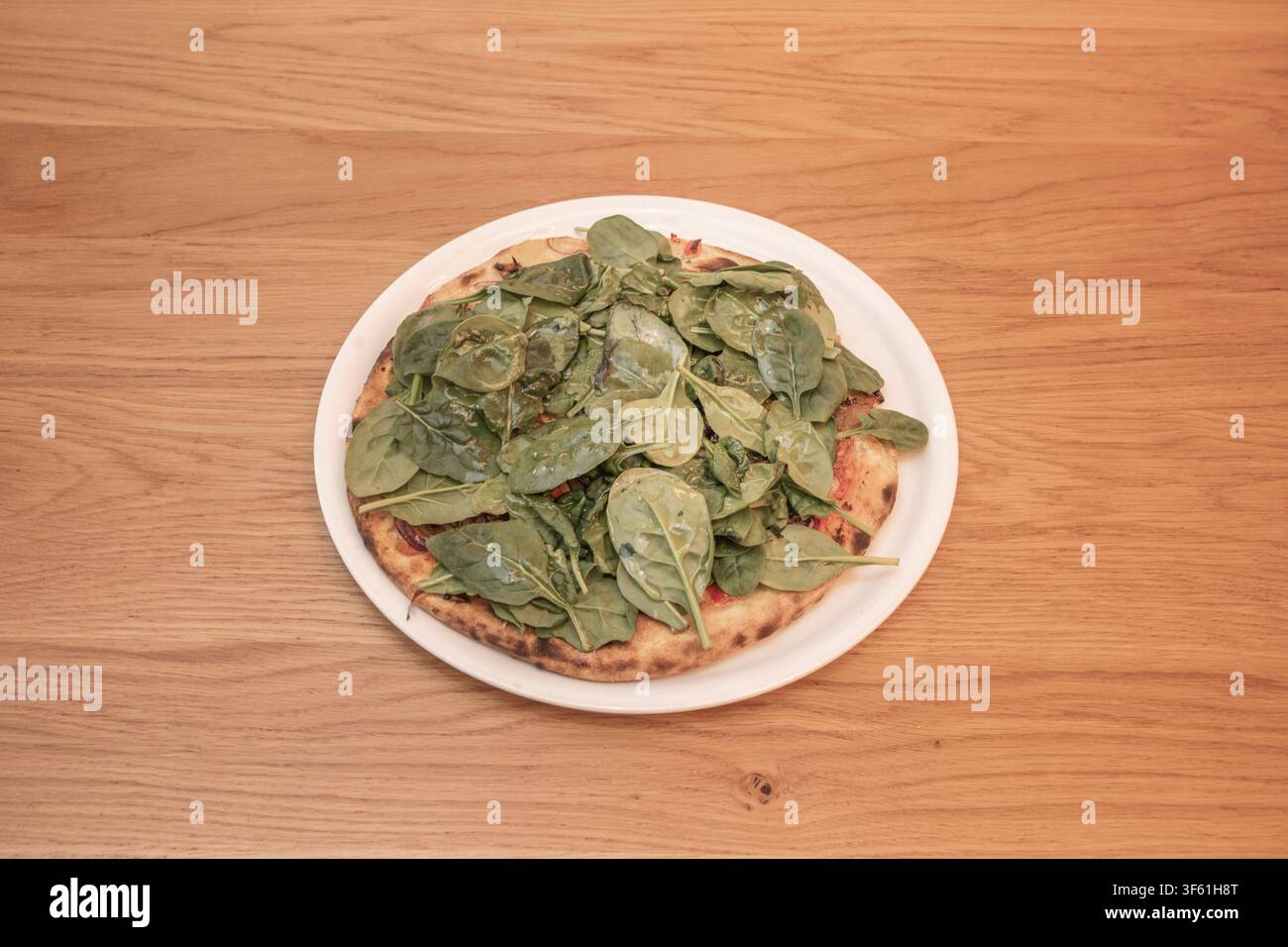 Pizza su pane azzimo ricoperta di foglie di spinaci freschi appassiti su piatto bianco sopra tavolo di legno Foto Stock