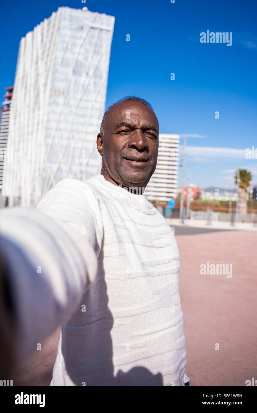 Uomo maturo che scatta un selfie sorridente davanti a un edificio moderno straordinario in una giornata soleggiata Foto Stock