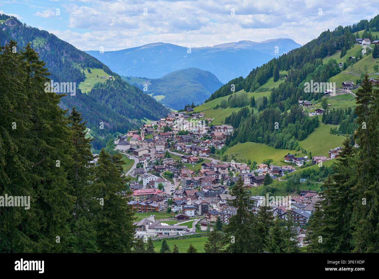 Vista aerea di un villaggio alpino incastonato in una valle montana con colline verdi e una foresta fitta. Foto Stock