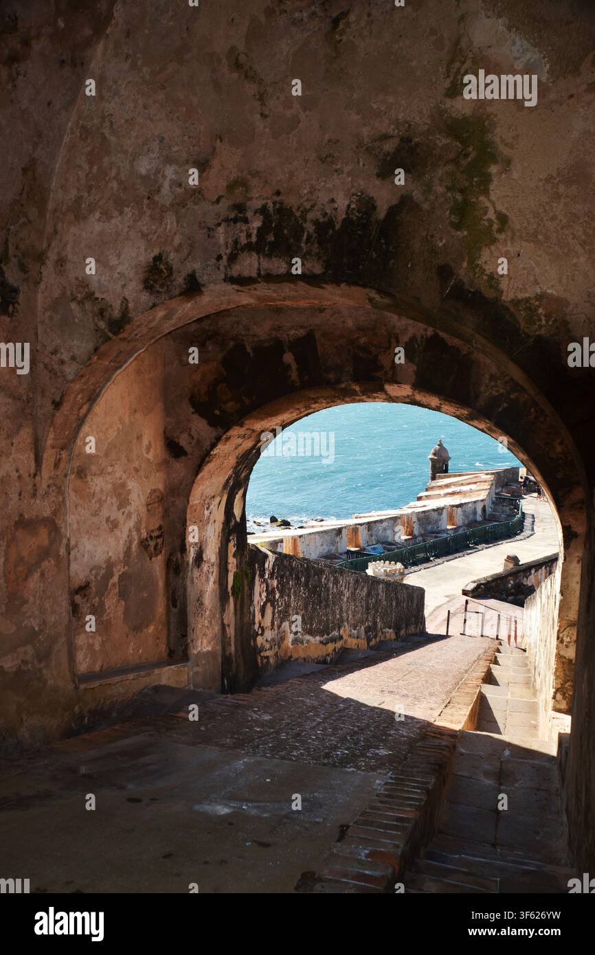 Un antico arco di pietra incornicia una vista costiera illuminata dal sole. Le mura erose mostrano l'età e una crescita delicata di muschio. Gradini di pietra ripidi conducono a acque blu brillanti. Lontano Foto Stock