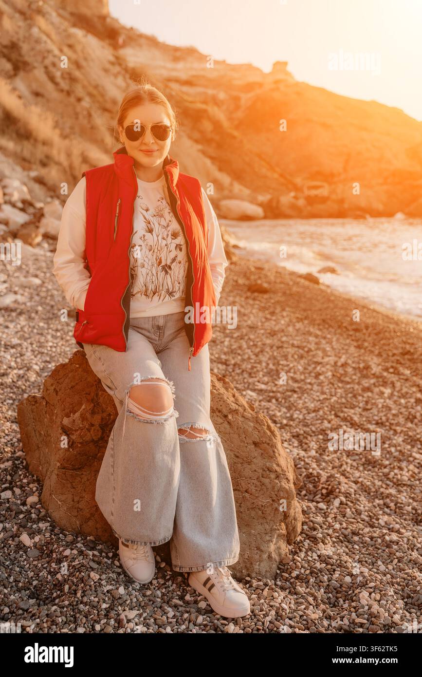 Donna in spiaggia al tramonto seduta su una roccia su una riva di ciottoli durante la calda luce dell'ora dorata Foto Stock
