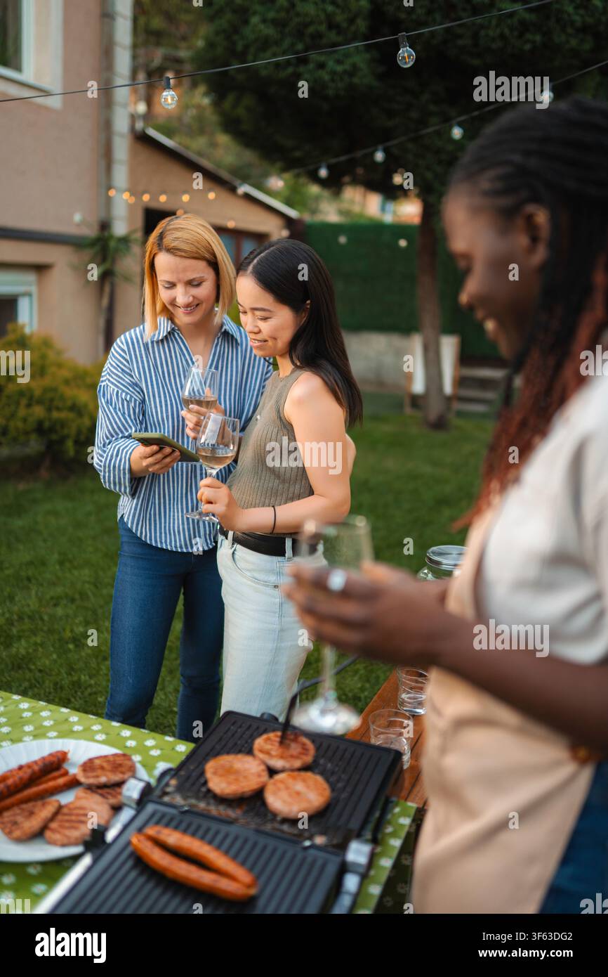 Diverse amiche donne che godono di un barbecue in giardino e usano gli smartphone Foto Stock
