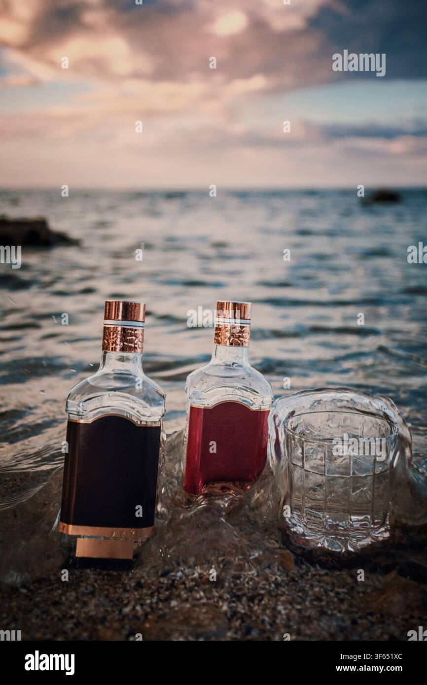 Due bottiglie di liquore con etichette vuote e vetro in piedi tra le onde del mare su una riva rocciosa al tramonto. Concetto di prodotto di lusso per alcolici con sfondo drammatico del cielo Foto Stock