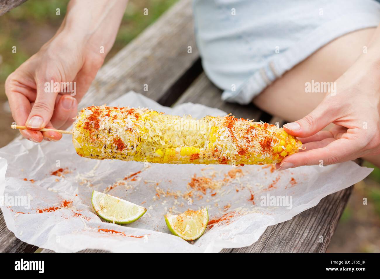 Mani tengono elote di mais messicano grigliato ricoperto di formaggio e polvere di chili su carta pergamena con spicchi di lime, ideale per il cibo da strada o l'estate Foto Stock