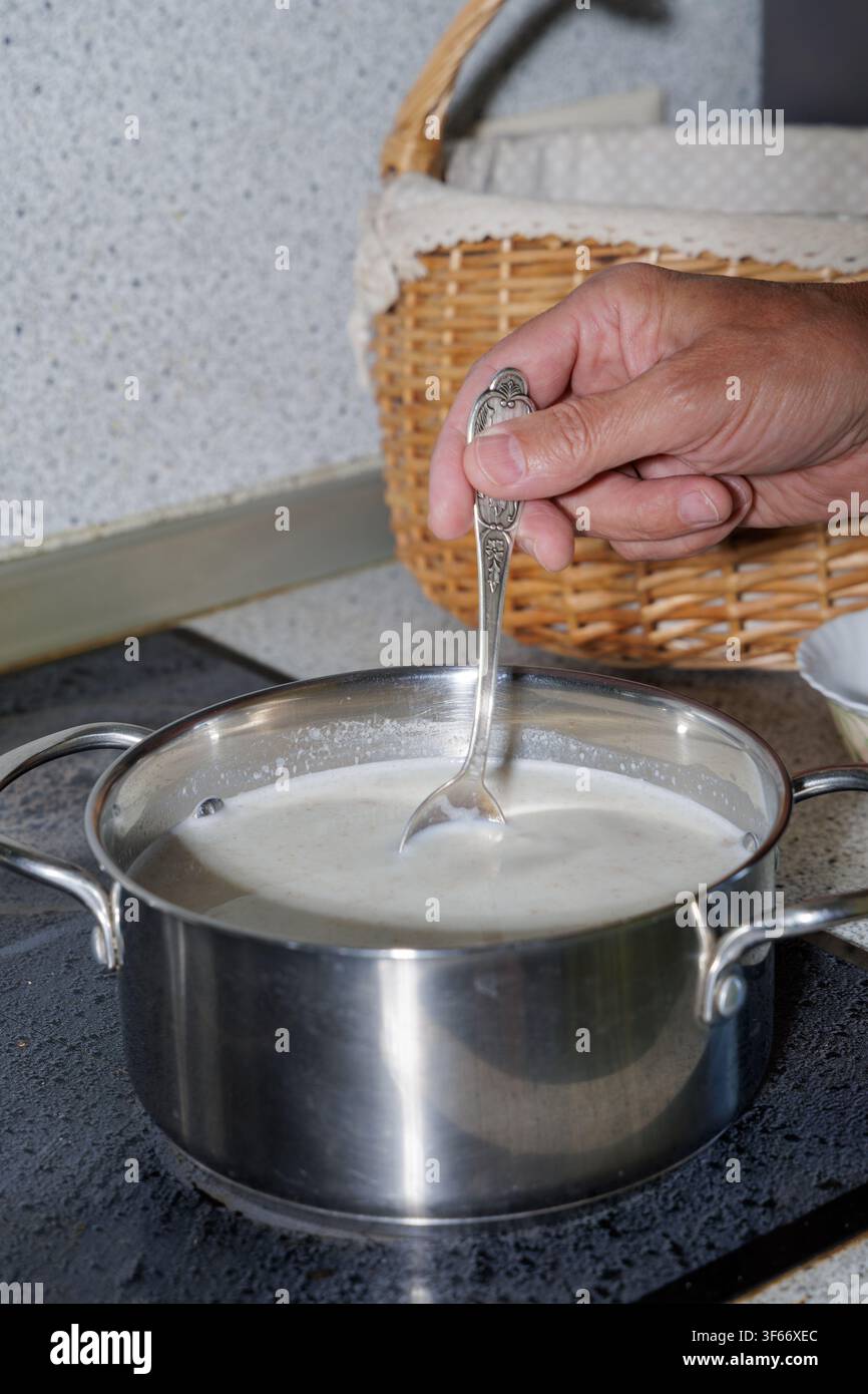 Prima piano di un uomo maturo concentrato che mescola un porridge di avena fatto in casa in una pentola di acciaio inossidabile sul fornello durante la sua routine mattutina a casa. Foto Stock