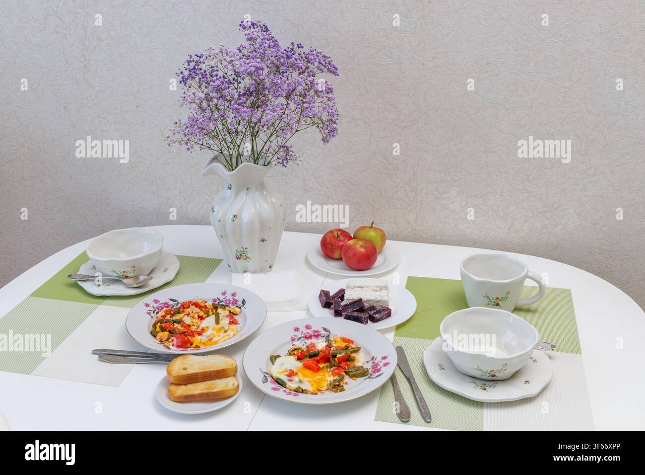 Vista ad angolo alto di un tavolo da cucina bianco apparecchiato con uova fritte, toast croccanti, farina d'avena, mele e fiori bellissimi per il pasto mattutino Foto Stock