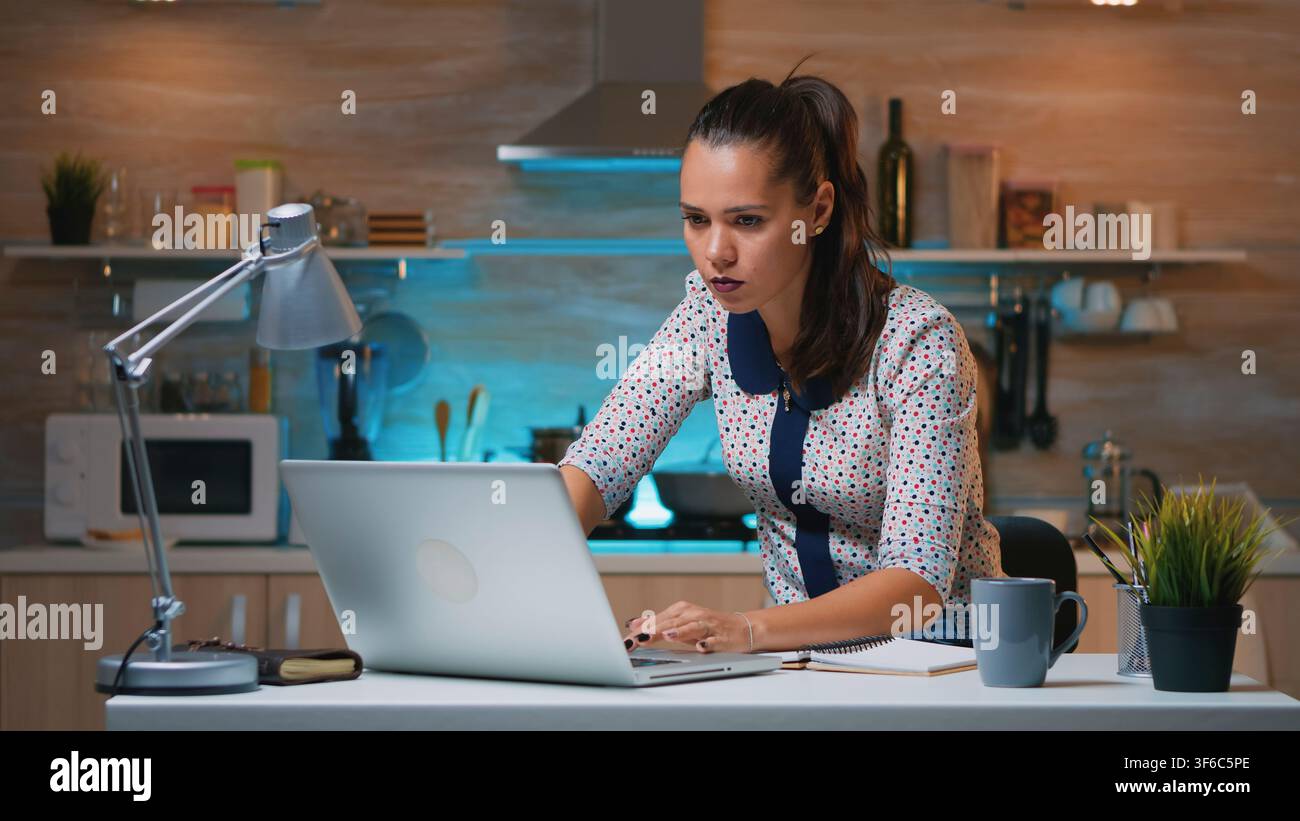 Donna scrive informazioni su un taccuino mentre consulta il suo laptop durante una sessione di lavoro tardo notturna concentrata dalla cucina. Foto Stock