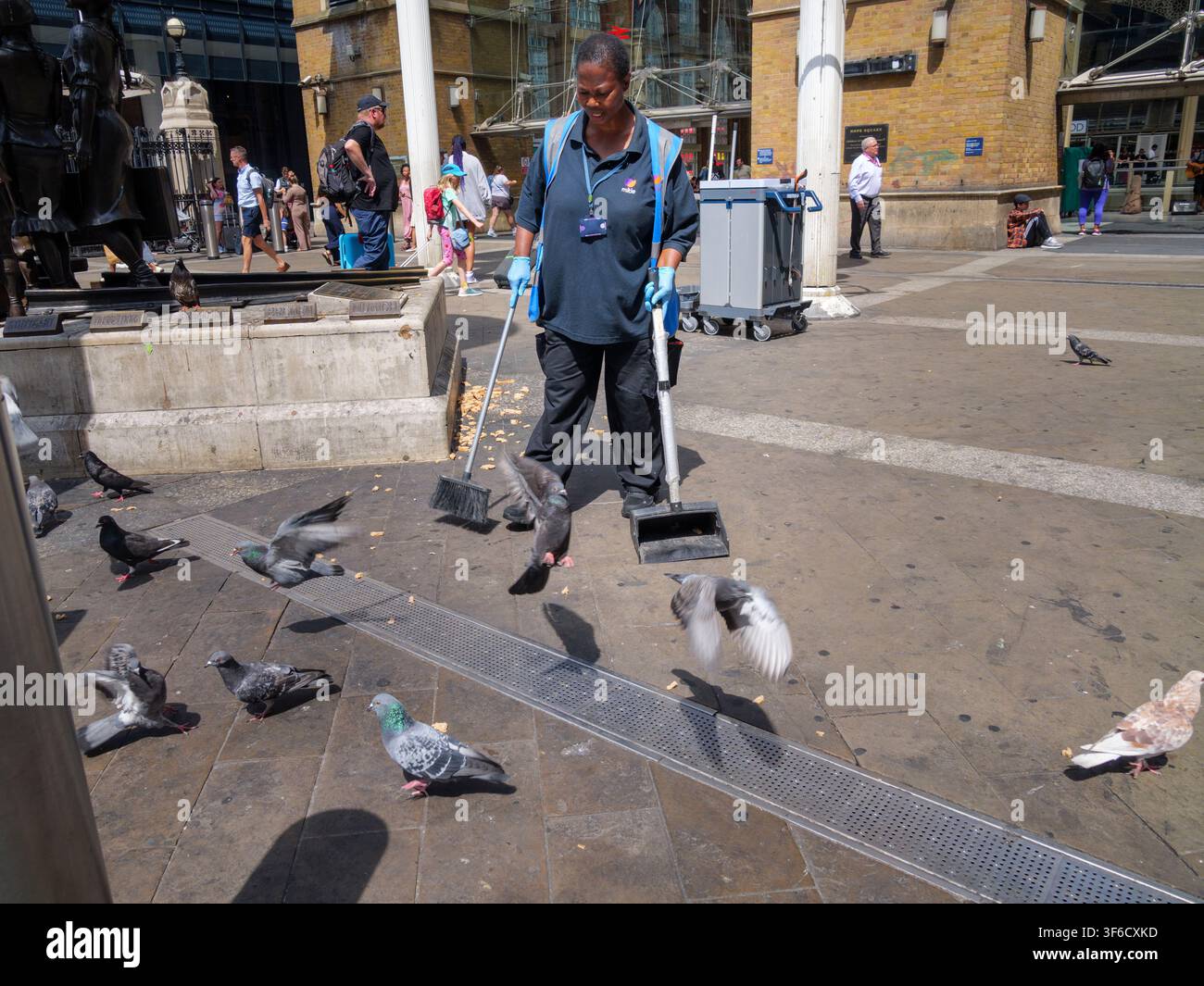 Un addetto alla manutenzione delle strutture impiegato da Mitie utilizza diligentemente una paletta per la polvere con manico lungo e una scopa per spazzare via le briciole di pane sparse sul marciapiede all'esterno dell'ingresso della stazione di Liverpool Street nella City di Londra, UK, mentre uno stormo di piccioni selvatici urbani scende intorno alla scena per mangiare rapidamente i resti rimanenti Foto Stock