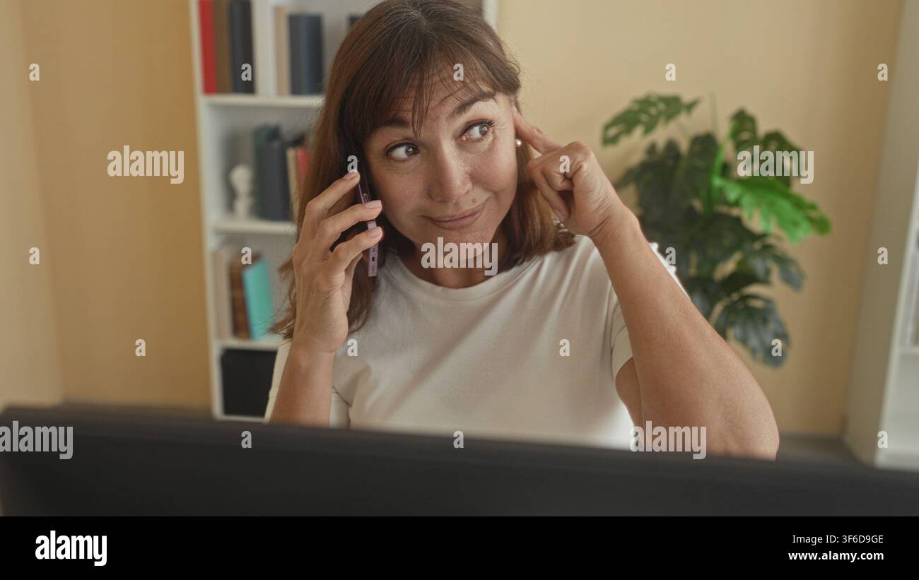 Una donna che parla con uno smartphone punta le dita verso un tempio in ufficio con lo schermo del computer visibile; messa a fuoco accurata. Foto Stock