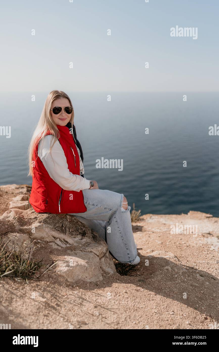 Donna seduta su una scogliera sul mare, con un giubbotto rosso e occhiali da sole, che gode della vista costiera con spazio di copia Foto Stock