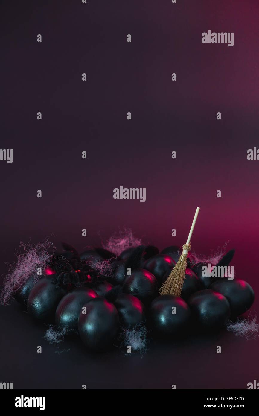Natura morta atmosferica di Halloween in studio con uova dipinte di nero, una piccola scopa da strega e piume bianche sparse su uno sfondo sfumato viola scuro Foto Stock