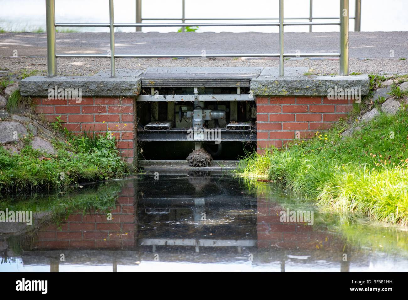 Piccola chiatta d'acqua con meccanismo metallico e muro di mattoni rossi che regola il livello dell'acqua in un canale del parco. Foto Stock