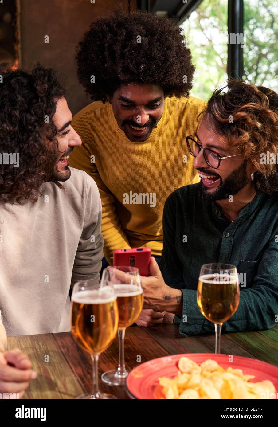 Un gruppo di amici in un bar che si diverte, beve birra, usa un telefono per guardare i social media. Foto Stock