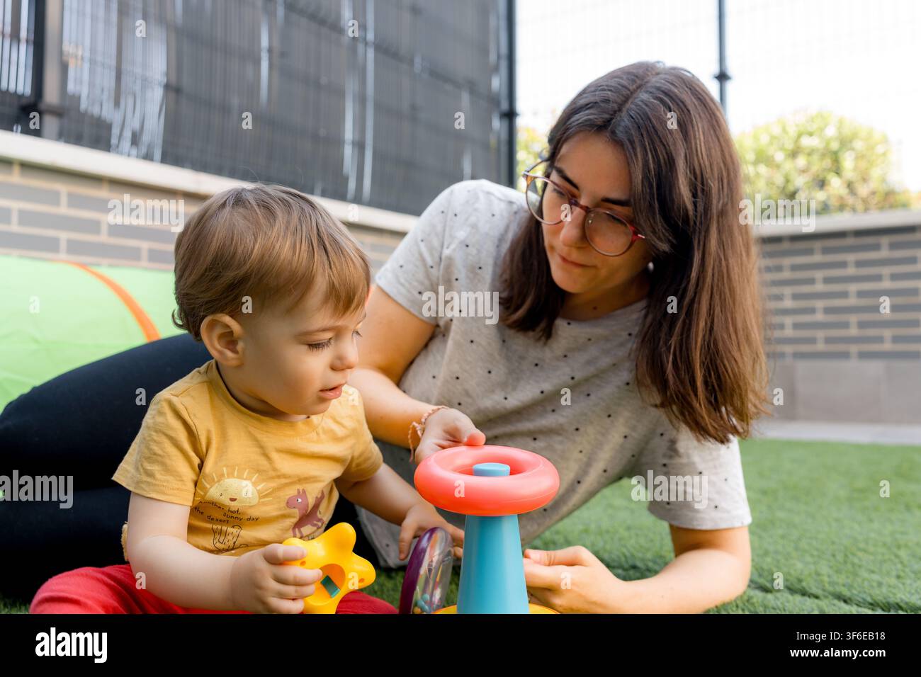 Madre con occhiali aiuta il suo bambino a esplorare un giocattolo colorato all'aperto su erba artificiale, creando un momento familiare caldo di apprendimento precoce, cura e gioco Foto Stock