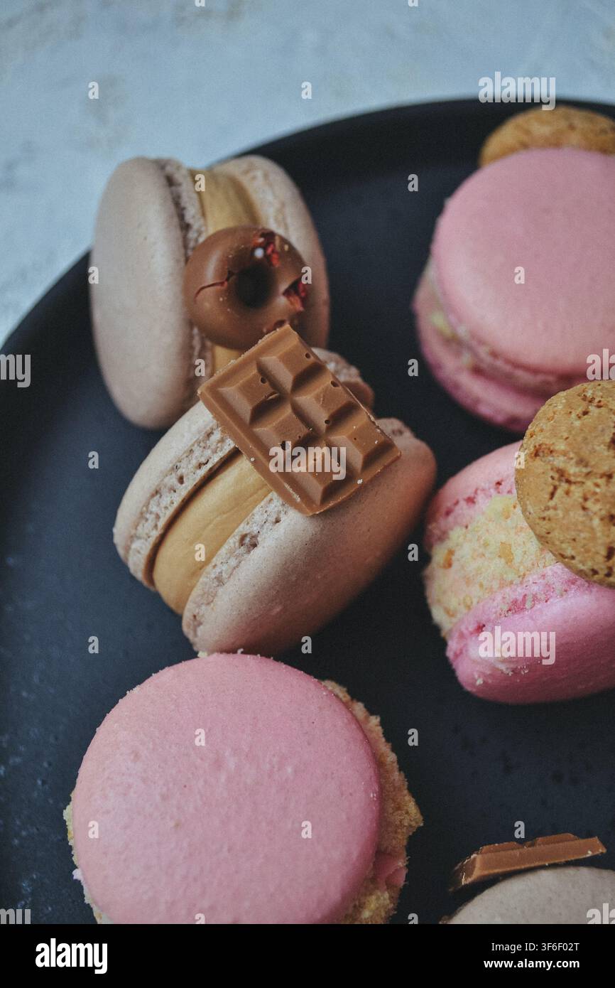 Macarons francesi assortiti decorati con mini barrette di cioccolato e biscotti serviti su un piatto nero Foto Stock