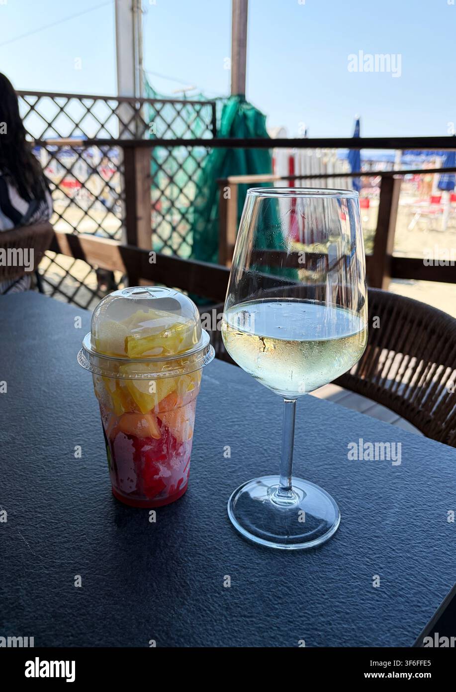 Bicchiere di vino bianco e coppa di frutta fresca su un tavolo del caffè sulla spiaggia con vista sul mare, che cattura un'atmosfera rilassante di vacanza estiva. Foto Stock