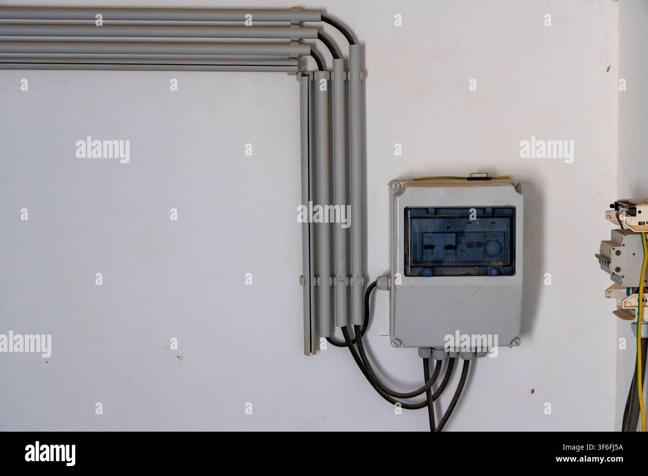 Conduiti elettrici grigi corrono lungo un muro bianco, portando a una scatola di distribuzione grigia con interruttori a carico visibili all'interno Foto Stock