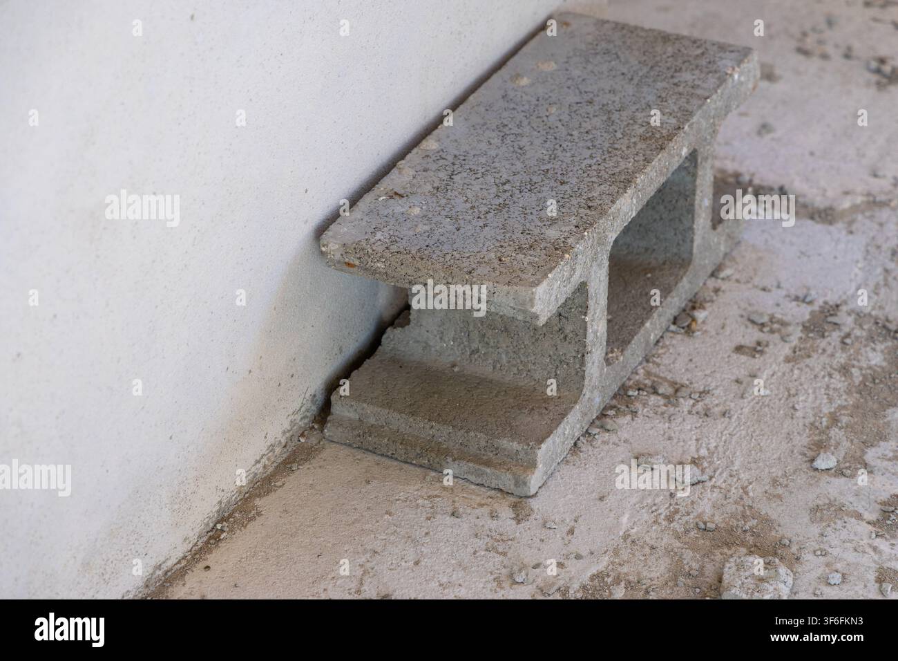 Un singolo blocco di cinder è appoggiato contro un muro ruvido, color crema chiaro, su una superficie concreta e ruvida con detriti sparsi Foto Stock